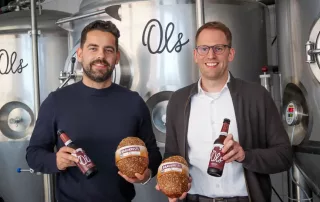 zwei personen stehen in der ols brauerei vor einem biertank. sie halten jeweils ein brot und eine flasche ols dunkel in der hand